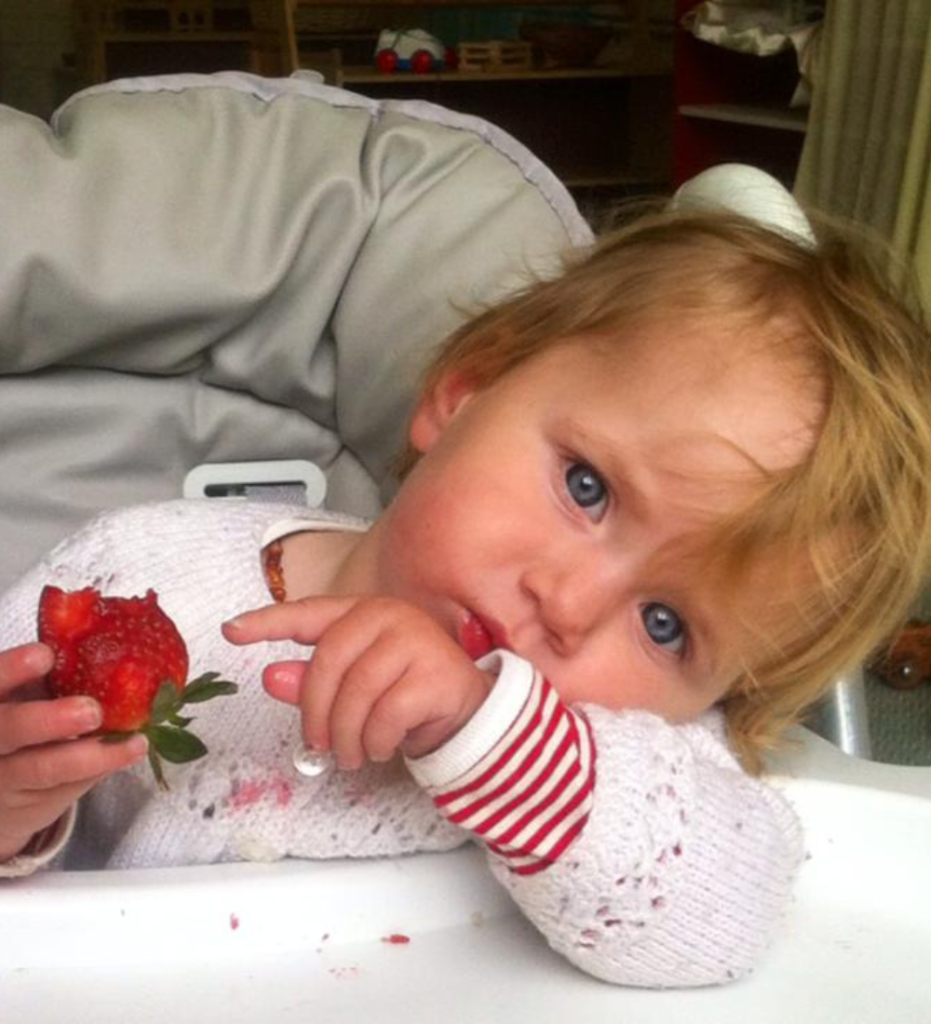 A infant sits on a high chair with her head cocked holding a strawberry. This is the kind of environment that begins a childs journey of science discovery and learning